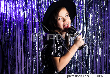 Portrait of a fashionable young girl in a black hat and a black dress singing with a microphone in 62409280