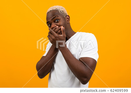 cute surprised african guy in a white t-shirt closes his mouth with his hands from surprise on a 62409599