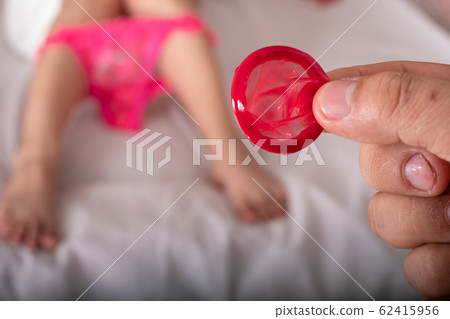 Man showing red condom at the beautiful woman with towel lies in bed Man showing red condom at the beautiful woman with towel lies in bed 62415956