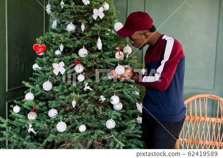 Portrait of an African-American hipster guy dressed in a red fleece sweater and red cap standing near the Christmas tree Portrait of an African-American hipster guy dressed in a red fleece sweater and red cap standing near the Christmas tree 62419589