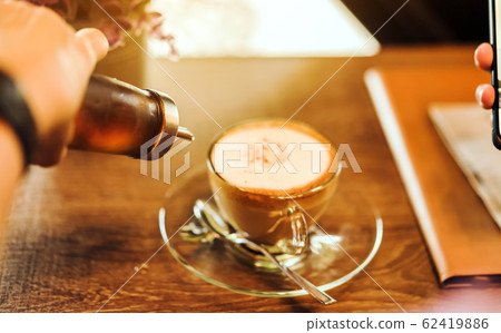 A woman poured the syrup into the saucer of the coffee cup while absent-mindedly playing the smartphone. Selective focus on the edge of the bottle 62419886