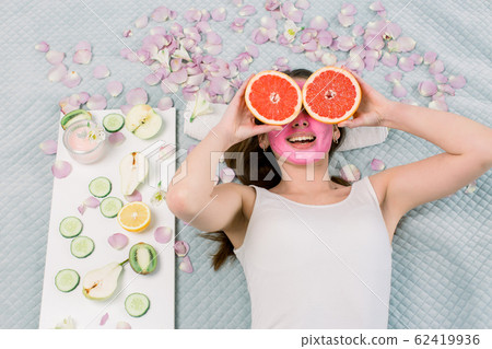 Young cheerful european woman with pink mud facial mask lies on bed and holds big grapefruits, covering her eyes. Healthy diet, skincare, facial treatment, cosmetology concept. 62419936