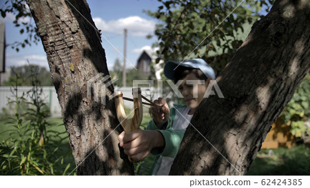 boy hides behind a tree and aims at someone with a slingshot boy hides behind a tree and aims at someone with a slingshot 62424385