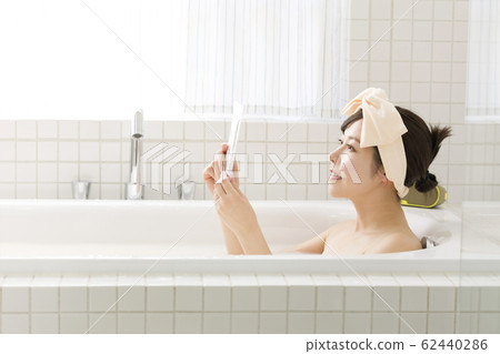 通過將智能手機放在浴室的防水盒中看智能手機的年輕女子 通過將智能手機放在浴室的防水盒中看智能手機的年輕女子 62440286