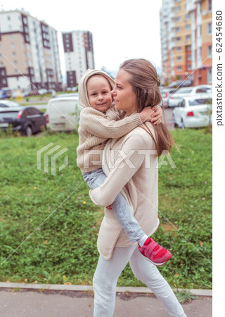 young family, woman, mother holds son in her arms, walks child, little boy 4-5 years old, in autumn in city, background of building. Emotions of love care and support. Casual warm clothes with hood. 62454680