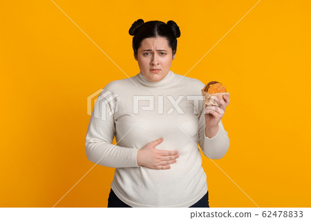 Stomachache. Girl Holding Burger And Touching Stomach Feeling Bad After Eating Junk Food Standing On Yellow Studio Background 62478833