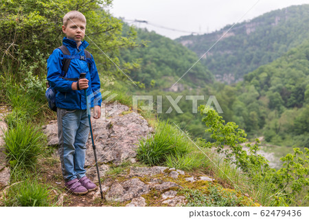 A boy with a backpack and trekking poles stands on a stone rock A boy with a backpack and trekking poles stands on a stone rock 62479436