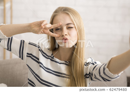 Cheerful girl taking selfie, showing duck face and peace gesture 62479480
