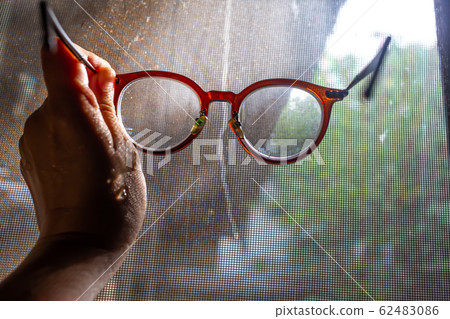 Woman's wet hand holding shortsighted or nearsighted eyeglasses with Dust and Dirty mosquito wire screen window, Close up shot, Selective focus 62483086