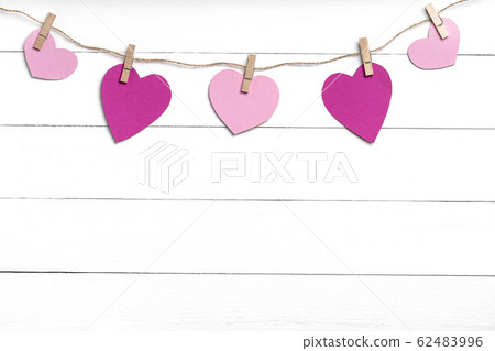 Clothespins with pink hearts hanging on rope behind white wooden surface. Clothespins with pink hearts hanging on rope behind white wooden surface. 62483996