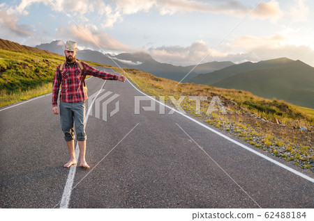 Stylish barefoot bearded male hitchhiker traveler in a hat and with a backpack walks along a suburban asphalt road in the mountains at sunset and stops the car with a hitch Stylish barefoot bearded male hitchhiker traveler in a hat and with a backpack walks along a suburban asphalt road in the mountains at sunset and stops the car with a hitch 62488184