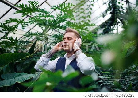 Young man with headphones standing in botanical garden. Young man with headphones standing in botanical garden. 62489223