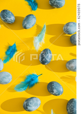 Blue textured colored Easter eggs and chicken bird feathers on a bright yellow holiday background backdrop. Spring holiday concept. Copyspace. Flat lay. Top view. Hard light. Long shadow. Blue textured colored Easter eggs and chicken bird feathers on a bright yellow holiday background backdrop. Spring holiday concept. Copyspace. Flat lay. Top view. Hard light. Long shadow. 62489978