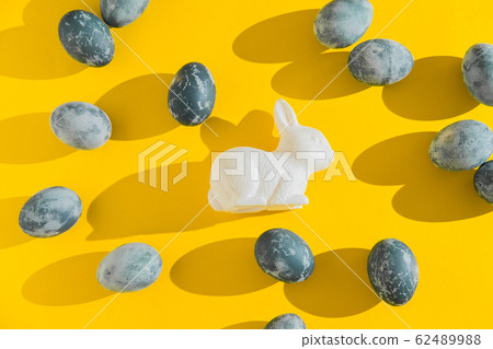 Blue textured colored Easter eggs and white plactic bunny on a bright yellow holiday background backdrop. Spring holiday concept. Copyspace. Flat lay. Top view. Hard light. Long shadow. 62489988