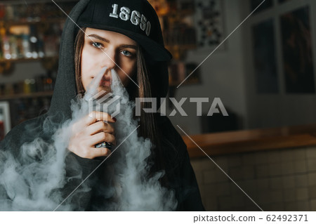 Young pretty woman in cap smoke an electronic cigarette at the vape shop. Hip-hop style. Closeup. Young pretty woman in cap smoke an electronic cigarette at the vape shop. Hip-hop style. Closeup. 62492371