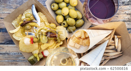 Tapas and pinchos, overhead panorama. Cheese, jamon and olives, gildas and potato chips, with glasses of red and white wine, on a wooden table in an outdoors cafe Tapas and pinchos, overhead panorama. Cheese, jamon and olives, gildas and potato chips, with glasses of red and white wine, on a wooden table in an outdoors cafe 62500816