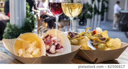 Tapas bar panorama. Cheese, ham and pinchos with red and white wine on a wooden table in an outdoors cafe Tapas bar panorama. Cheese, ham and pinchos with red and white wine on a wooden table in an outdoors cafe 62500817