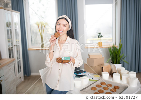 Slender woman standing with a saucer of cookies, one in hand. Slender woman standing with a saucer of cookies, one in hand. 62501727