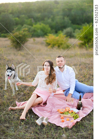 Young woman and man sitting on pink plaid in steppe near dog husky. Young woman and man sitting on pink plaid in steppe near dog husky. 62502583