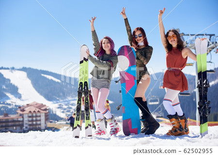 Girls in summer clothing posing in snow with snowboard and skis on background of winter ski resort. 62502955