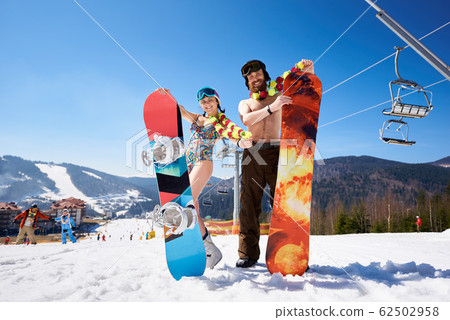 Girl in bra and skirt and bearded man with snowboards on background of winter mountains resort. Girl in bra and skirt and bearded man with snowboards on background of winter mountains resort. 62502958