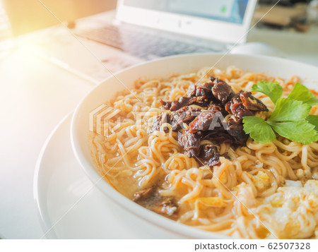 Asia instant noodle boiled with egg and Tom Yum Kung soup, decorated by fried baby clams with chilli and celery leaves, in white bowl and dish, on table and computer or notebook with orange sunshine. Asia instant noodle boiled with egg and Tom Yum Kung soup, decorated by fried baby clams with chilli and celery leaves, in white bowl and dish, on table and computer or notebook with orange sunshine. 62507328