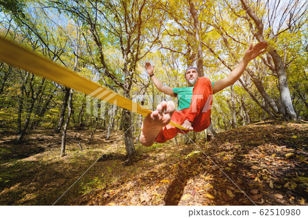Wide angle male tightrope walker balancing while sitting barefoot on slackline in autumn forest. The concept of outdoor sports and active life of people aged 62510980
