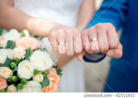 Hands of the newlyweds bride and groom with gold wedding rings close-up on a background of a wedding bouquet of roses 62511013