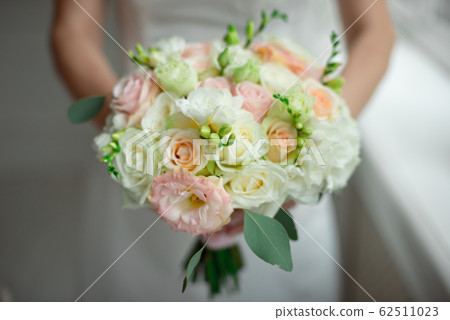 Close-up groom in a suit and the bride in a white dress are standing and holding a bouquet of peach roses, eustomas and flowers and greenery 62511023