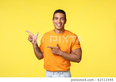 Cheerful modern man with tattooed arm, pointing left as promoting online store, invite guest join party, smiling joyfully, recommend try something, give advice what place visit, yellow background 62511444