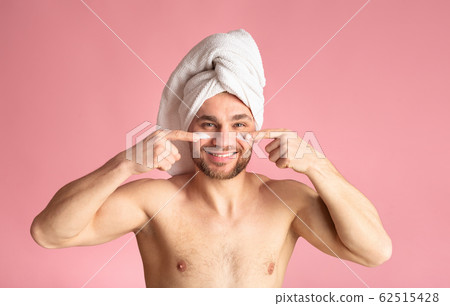 Cute young guy after a shower, white towel on head, smears cream on face Cute young guy after a shower, white towel on head, smears cream on face 62515428