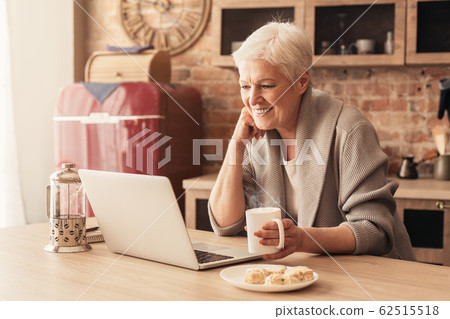 Cheerful senior woman using laptop in kitchen and drinking coffee Cheerful senior woman using laptop in kitchen and drinking coffee 62515518
