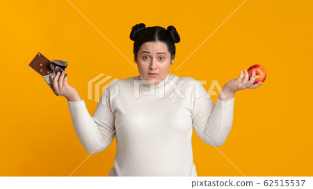 Puzzled Overweight Woman Holding Chocolate Bar And Apple In Hands Puzzled Overweight Woman Holding Chocolate Bar And Apple In Hands 62515537