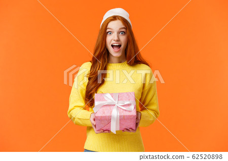 Surprised and excited happy girl unwrapping gifts on christmas eve, new year celebration, smiling amused and thrilled, holding wrapped box present, wearing winter beanie, orange background 62520898