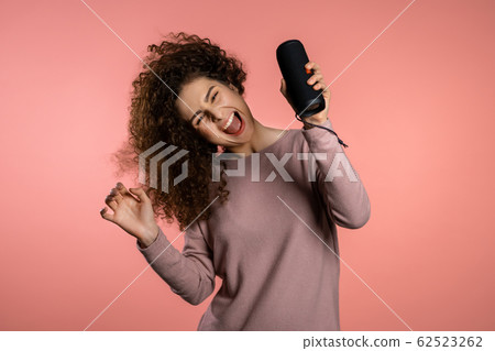 Cute woman with curly hair smiling, dancing with wireless portable speaker in studio on pink background. Music, dance concept 62523262