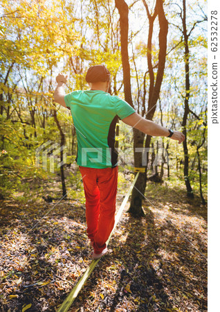 Wide angle male tightrope walker balancing barefoot on slackline in autumn forest. The concept of outdoor sports and active life of people aged 62532278