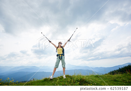 Woman hiker hiking on grassy hill, wearing backpack, using trekking sticks in the mountains 62539187