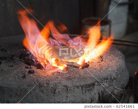 silver plate heating by craftsman using old fashioned appropriate fireplace with yellow orange flame from firewood blowing with manual hand blower in a silversmith art and crafts shop in LAOS 62541661