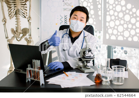 Happy Asian man scientist in white coat, mask and blue gloves, types on laptop the results of research, and shows his thumb up. Synthesis of new drugs, cancer treatment, medicine, biochemistry concept 62544664
