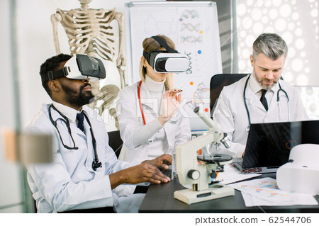 team of multiethnic scientists, African man, Caucasian woman and man, using virtual reality glasses for research in laboratory. Developmetn of new medicine, vaccine, substance team of multiethnic scientists, African man, Caucasian woman and man, using virtual reality glasses for research in laboratory. Developmetn of new medicine, vaccine, substance 62544706