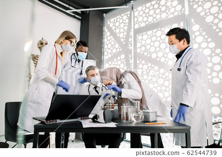 Team of scientists doctors biochemists working with microscope, doing research scientific experiments in laboratory. Muslim woman is looking through microscope and discussing with colleagues 62544739