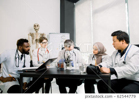 Team of multiethnic medical research scientists working in modern laboratory on a new generation drug or vaccine. Man in medical mask is looking in microscope while his colleagues making notes 62544779