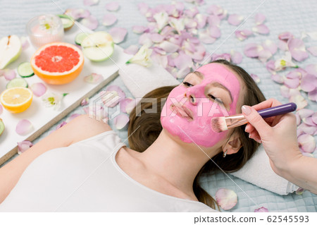 Close-up portrait of beautiful girl applying pink mud facial mask. Spa Woman applying Facial clay Mask. Beauty Treatments. Close-up portrait of beautiful girl applying pink mud facial mask. Spa Woman applying Facial clay Mask. Beauty Treatments. 62545593