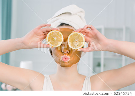 Emotional young woman in white towel on head and with facial mask and halves of ripe lemon on white background. Photo of woman receiving spa treatments. Beauty and Skin care concept 62545734