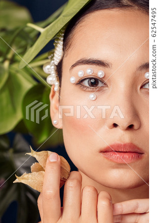 Freestyle. Young woman eyes decorated with perls standing isolated on dark blue with plants looking camera curious close-up 62547495
