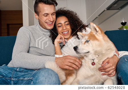 Romantic Date. Young multiethnic couple at home sitting on sofa playing with dog laughing cheerful Romantic Date. Young multiethnic couple at home sitting on sofa playing with dog laughing cheerful 62548028