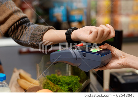 Daily Shopping. Woman standing at the supermarket cashier counter paying for purchases contactless with smartwatch close-up 62548045