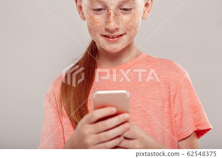 Inclusive Beauty. Girl with freckles standing isolated on grey using app on smartphone smilign concentrated close-up Inclusive Beauty. Girl with freckles standing isolated on grey using app on smartphone smilign concentrated close-up 62548375