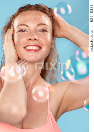 Summer Freestyle. Young woman with freckles standing isolated on blue soap bubbles touching hair posing cheerful Summer Freestyle. Young woman with freckles standing isolated on blue soap bubbles touching hair posing cheerful 62548381
