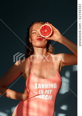 Summer Freestyle. Young woman with freckles and wet hair standing isolated on blue covering eye with grapefruit half posing grimace silly Summer Freestyle. Young woman with freckles and wet hair standing isolated on blue covering eye with grapefruit half posing grimace silly 62548383